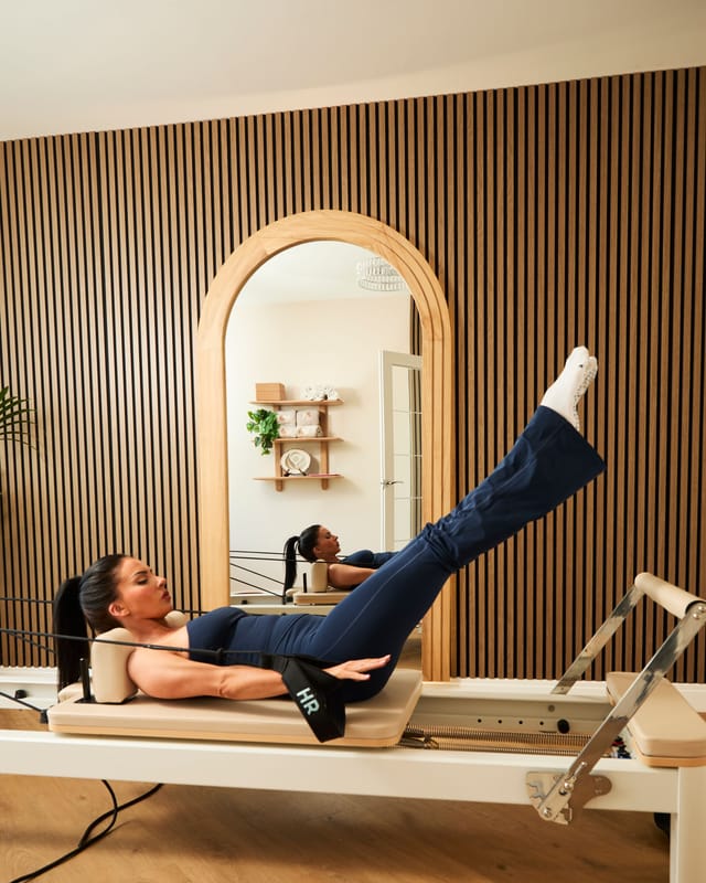 A person lying on a Pilates reformer with their leg extended upward, reflected in an arched mirror on a fluted wooden wall.