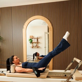 A person lying on a Pilates reformer with their leg extended upward, reflected in an arched mirror on a fluted wooden wall.