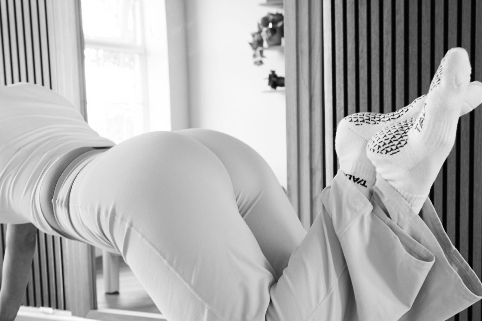 A person lies on a Pilates reformer with their head and shoulders in view as they perform an exercise in a bright studio with floor-to-ceiling curtains.